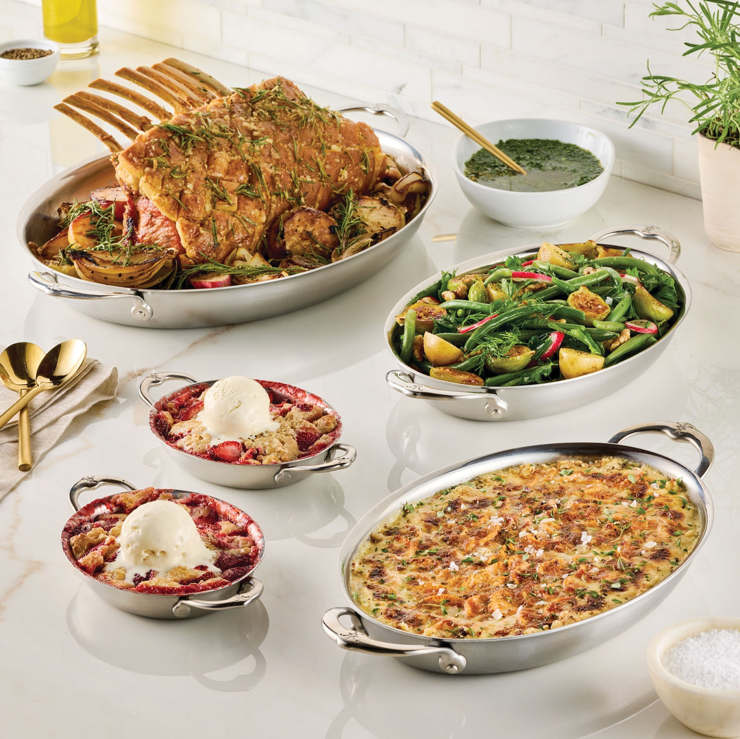 A table displays a spread of dishes including a rack of seasoned roasted meat with vegetables, a green salad with croutons, a baked dish with herbs, and two bowls of dessert topped with ice cream. A small bowl of sauce is in the background.