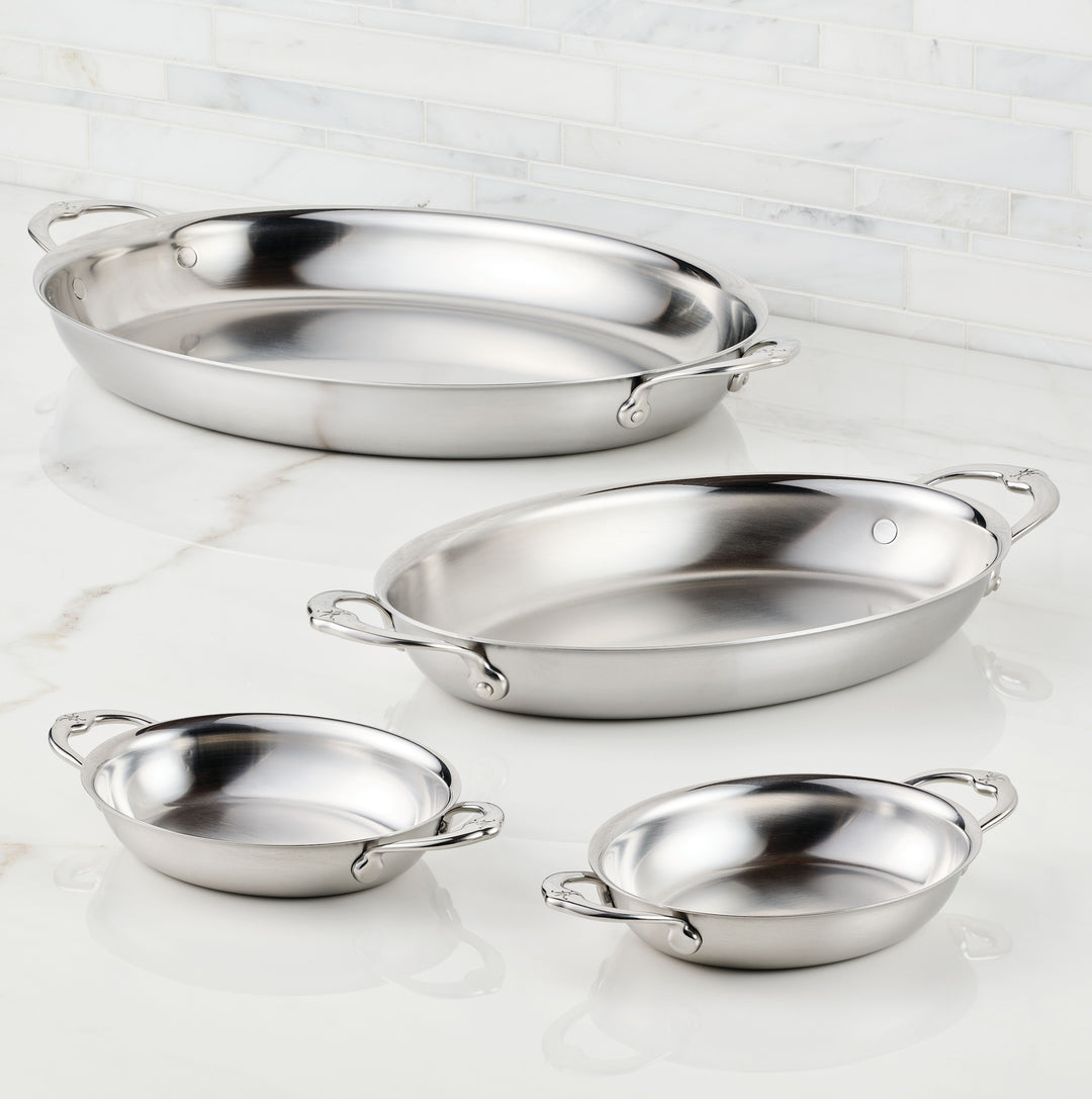 Four stainless steel pans with handles are placed on a marble countertop. The pans vary in size, with two larger oval pans at the back and two smaller round pans in the front. A white tiled wall forms the backdrop.