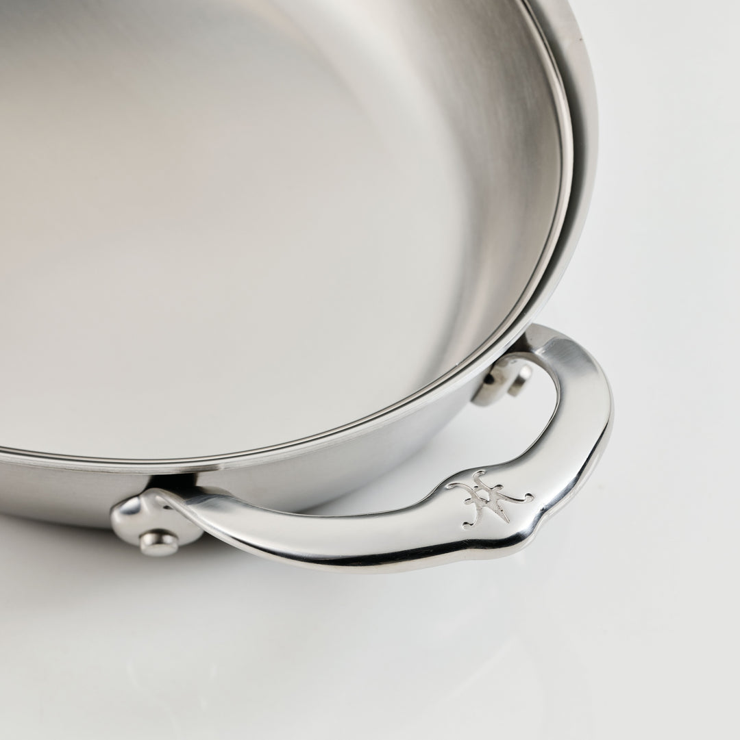 A close-up of a stainless steel casserole dish focusing on the handle. The handle has a decorative engraving and is attached with two rivets. The finish is polished, reflecting light softly, and the interior of the dish is smooth and shiny.