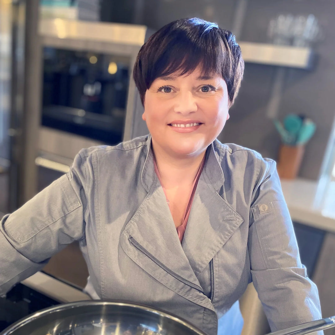 A person with short dark hair, wearing a grey chefs jacket, smiles while standing in a kitchen. They are positioned behind a counter with a metal mixing bowl in the foreground. The kitchen has blurred shelves and utensils in the background.