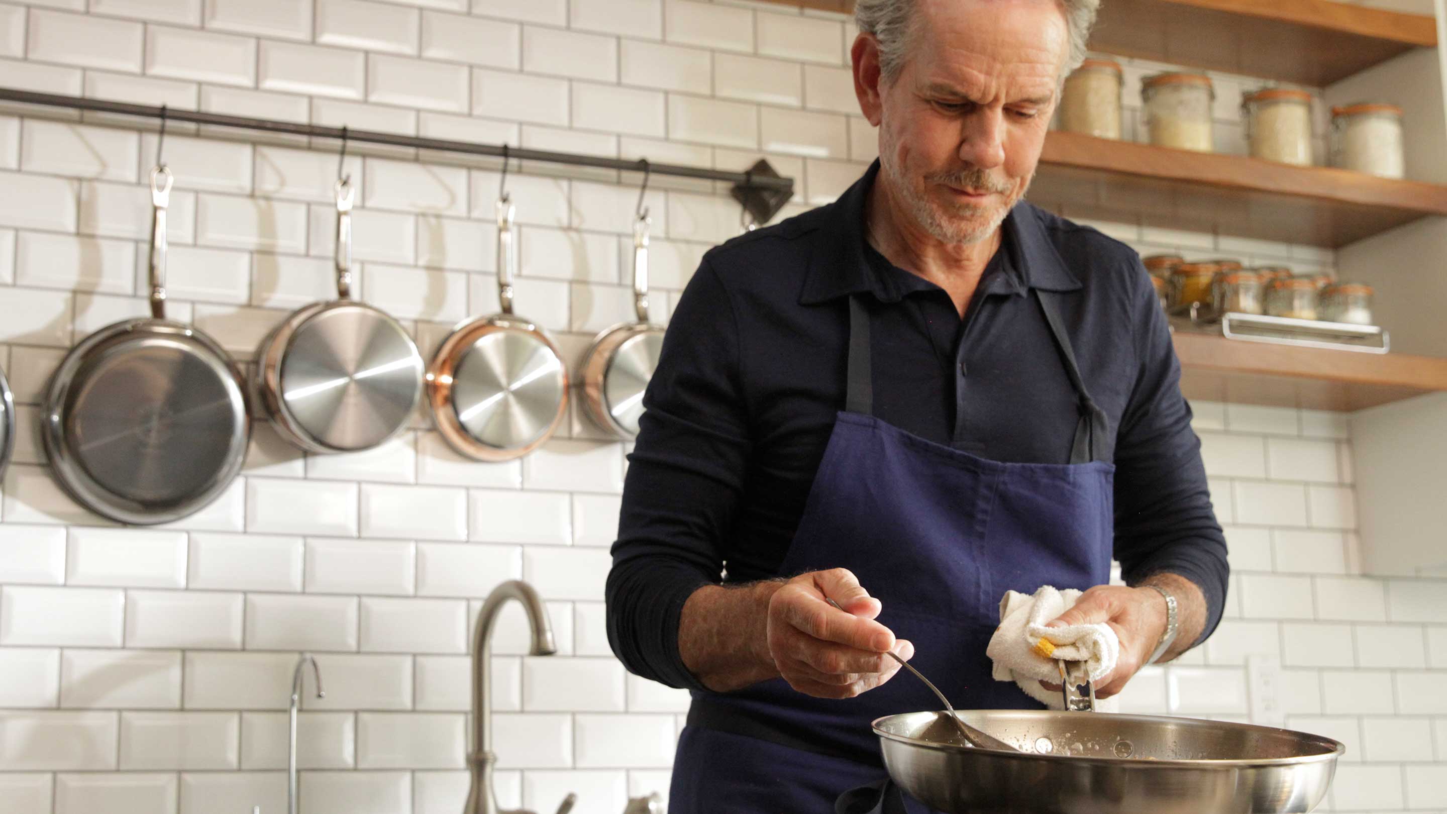 A man with grey hair is cooking in a kitchen, wearing a blue apron over a dark shirt. He is stirring a pan on the stove. The background features white subway tiles, a shelf with jars, and a hanging rack with pots and pans.