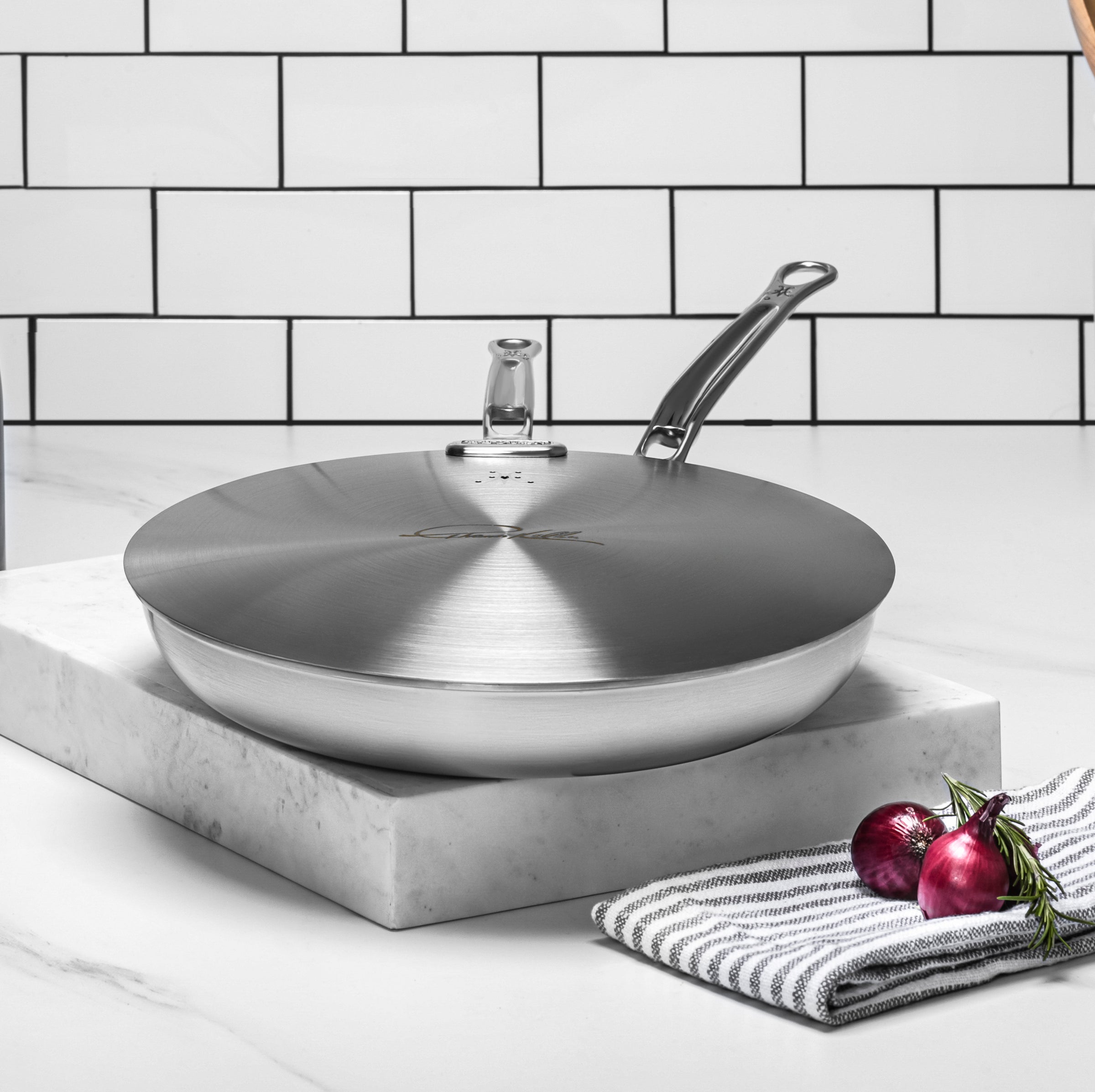 A stainless steel frying pan with a lid, featuring a sleek handle, sits on a white marble surface. Nearby, a striped cloth and a pair of purple onions serve as accents. The backdrop consists of white subway tiles.