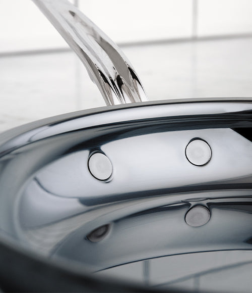 Close-up of a shiny chrome kitchen sink faucet extending over a polished, curved stainless steel sink. The sink surface reflects light, showing circular overflow holes and the surrounding countertop.