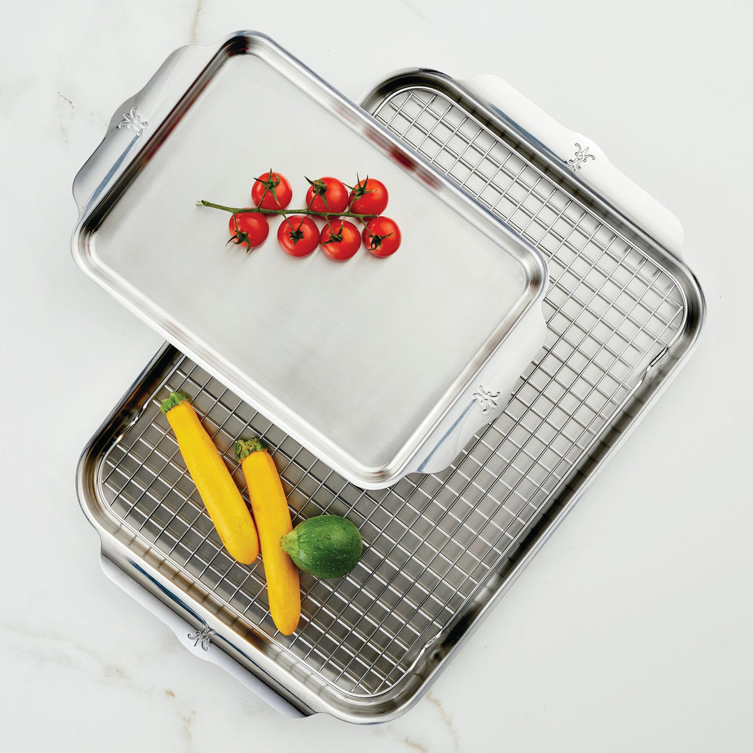 Two stainless steel baking trays are shown. The top tray has cherry tomatoes, and the bottom tray has yellow zucchinis and a green squash with a cooling rack. The background is a white surface.