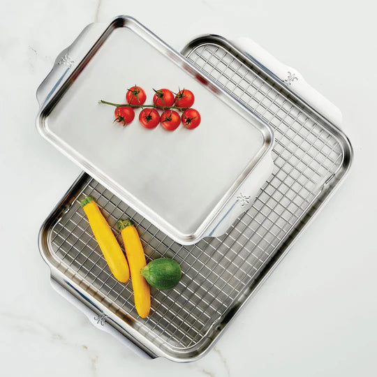 Two metal baking sheets are arranged on a light surface. The top sheet holds a cluster of cherry tomatoes, and the larger bottom sheet contains a metal cooling rack with two yellow squash and one green zucchini.