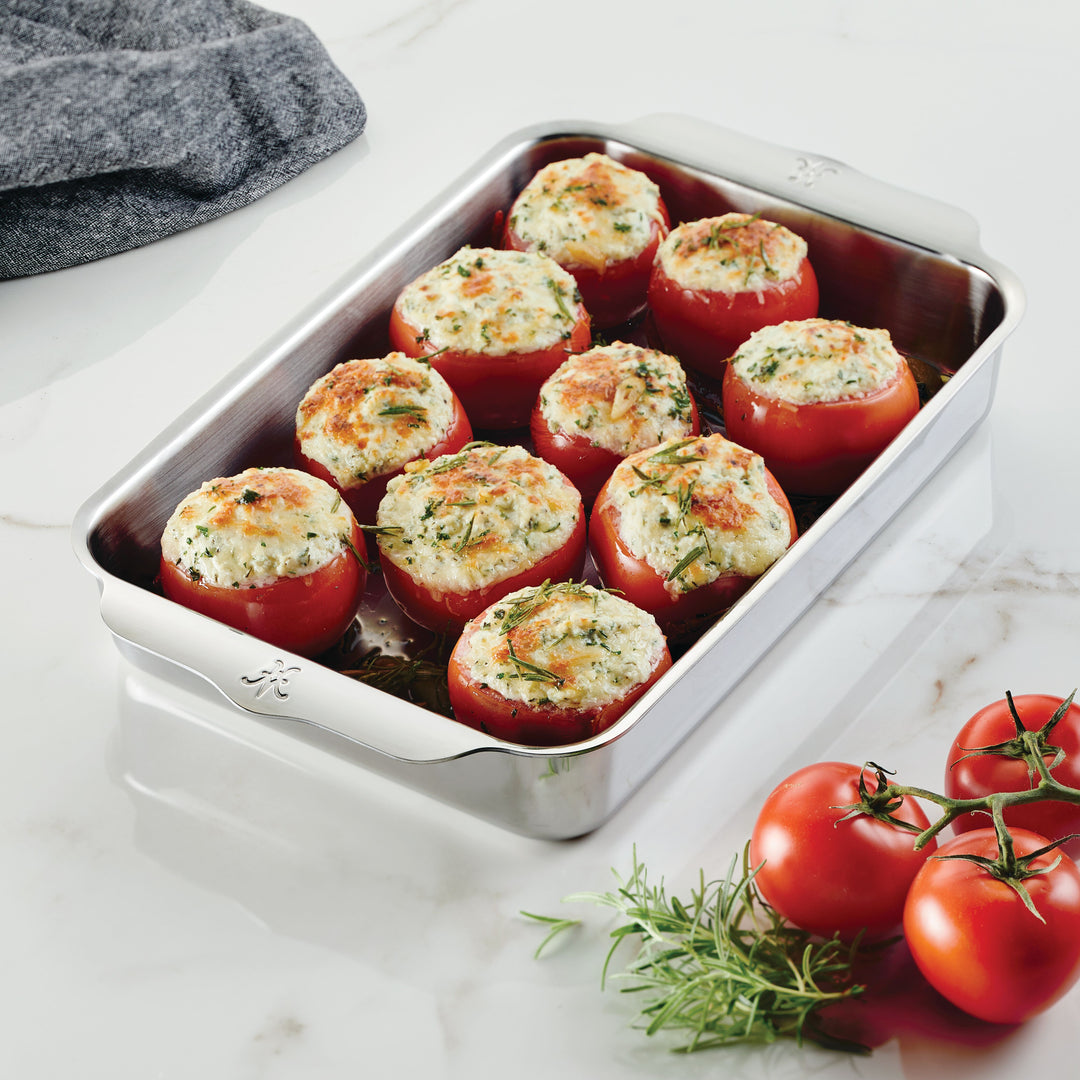 A stainless steel baking dish containing baked stuffed tomatoes on a marble countertop. The tomatoes are filled with a mixture topped with herbs and cheese. Fresh twig of rosemary and a bunch of tomatoes are placed beside the dish.
