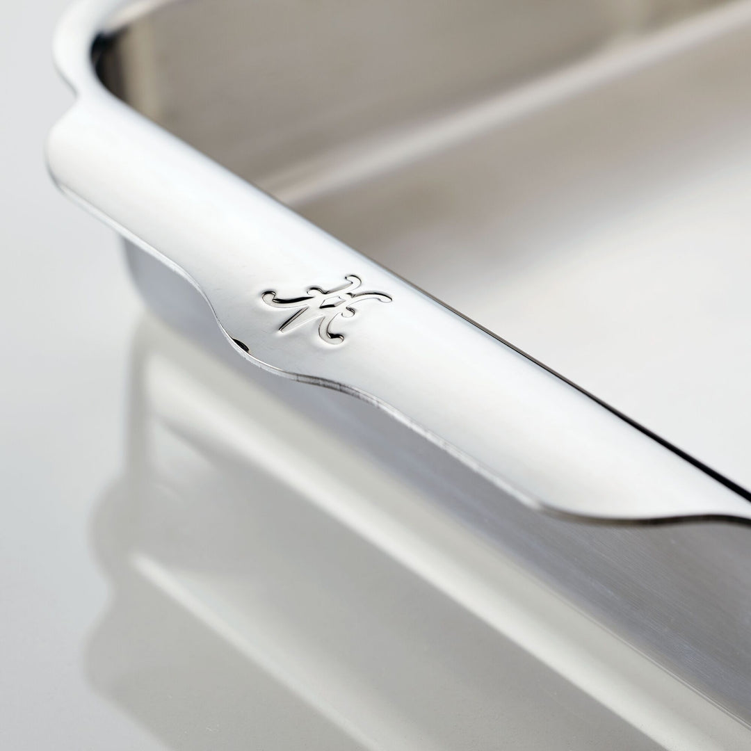 Close-up of a metal baking tray on a reflective surface. The edge of the tray features a decorative engraving of the letter A in a cursive style. The trays shiny surface is highlighted in the lighting.