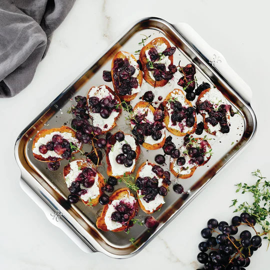A silver tray holds toasted bread slices topped with creamy cheese, roasted grapes, and garnished with thyme. A few grapes and thyme sprigs are scattered beside the tray on a white surface. A gray linen cloth is also visible in the corner.
