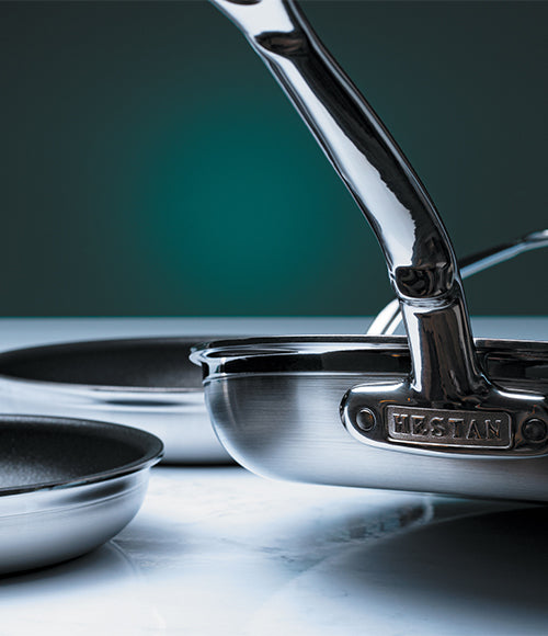Close-up of stainless steel cookware against a dark background. The focus is on the polished handle and the brand name HESTAN engraved on a metal piece. Two pans with dark interiors are partially visible on a white marble surface.