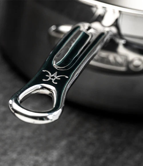 Close-up of a metal pot handle with a polished finish, featuring an intricate design resembling a logo or symbol. The background is blurred, highlighting the handles sleek and reflective surface.