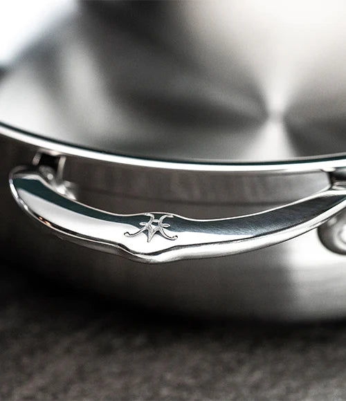 Close-up of a stainless steel saucepan handle with a logo engraved on it. The handle has a polished finish, and the background shows part of the saucepans lid. The surface beneath is a textured gray material.
