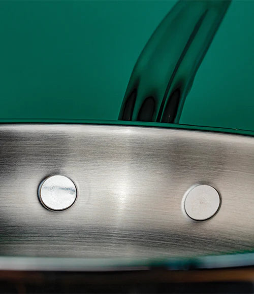 Close-up of a stainless steel pan with two rivets on the side. The background is a solid teal color. The pans edge and handle are visible, showcasing the metals smooth, polished surface.