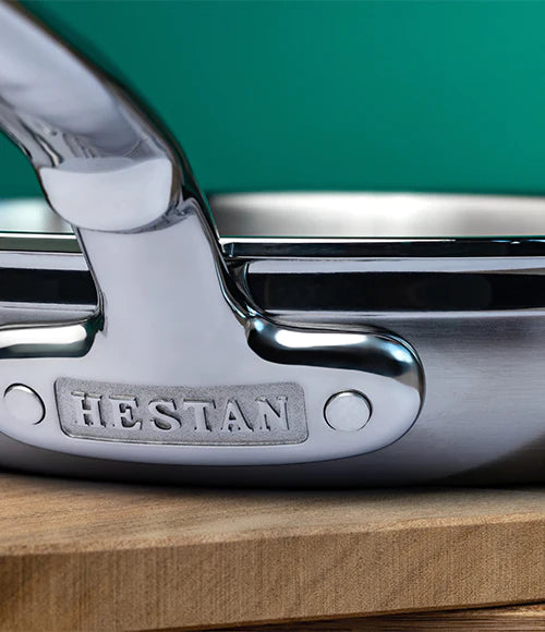Close-up of a stainless steel pan handle with the brand name HESTAN embossed on the side. The pan rests on a wooden surface against a green background.