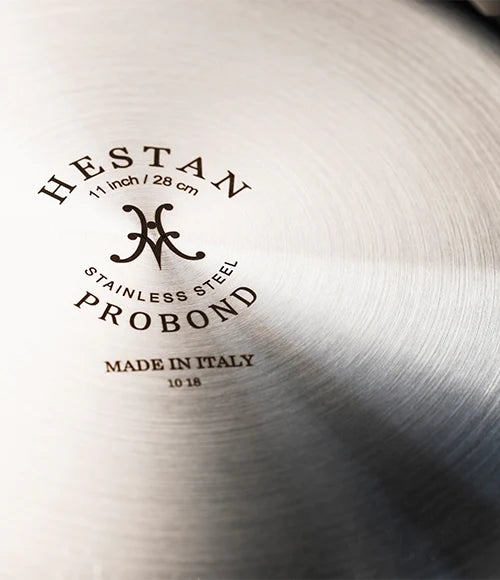 Close-up of the bottom of a stainless steel pan. The text reads HESTAN PROBOND, 11 inch / 28 cm, Stainless Steel, Made in Italy. The surface has a brushed metal texture.