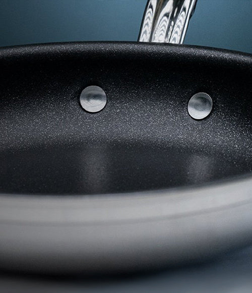 Close-up of a black non-stick frying pan with a silver handle. The interior surface is smooth with visible rivets securing the handle. The background is a soft, blurred blue.