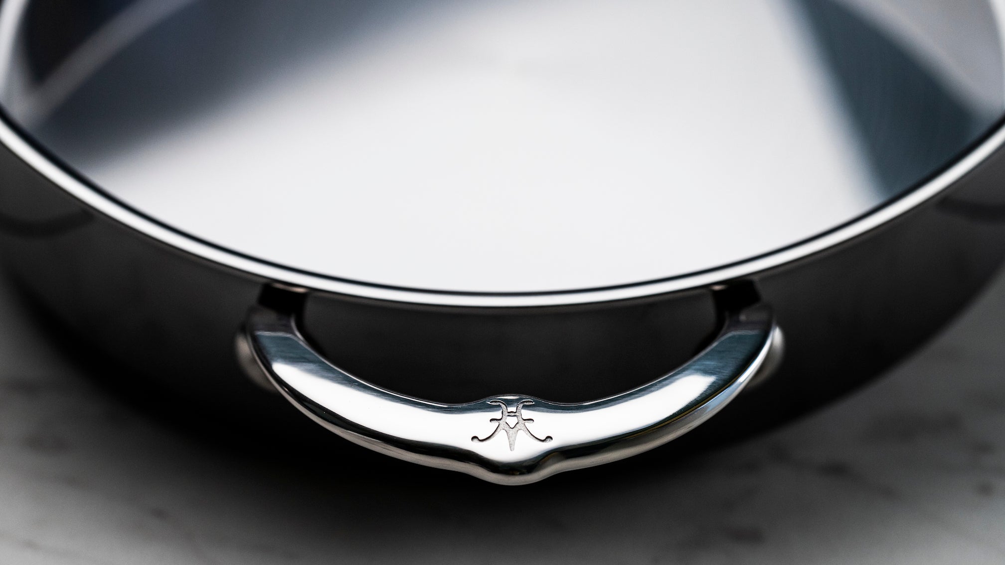 Close-up of a stainless steel pot with a polished handle, featuring a decorative emblem. The inside of the pot is smooth and reflective. The background is blurred, focusing attention on the pots design details.