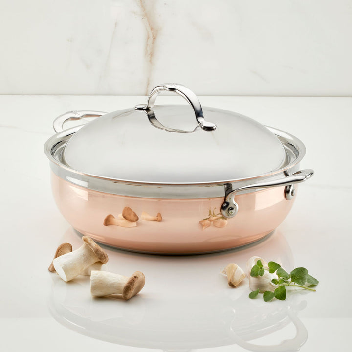 A CopperBond Copper Induction Dutch Oven, 5-Quart with a lid sits on marble. Nearby are three peeled garlic cloves, three mushrooms, and a small herb sprig against a subtly marbled white background.