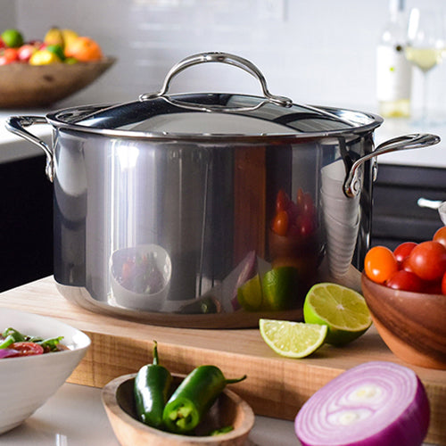 A NanoBond Titanium Stockpot with a lid rests on a wooden countertop, surrounded by fresh tomatoes, limes, jalapeños, red onion, and a salad bowl in a bright, modern kitchen.