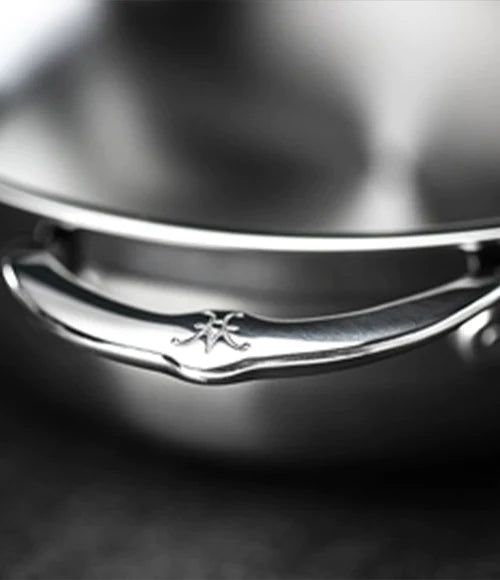 Close-up image of a stainless steel pot handle with an engraved logo. The handle is attached to the smooth, reflective surface of the pot. The overall tone is metallic and sleek, set against a dark background.