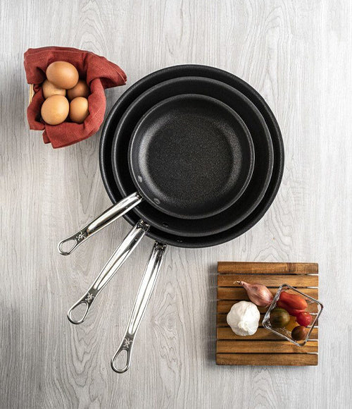 Three black frying pans with metal handles are stacked on a white wooden surface. Nearby, there is a red cloth holding eggs, and a small wooden board with ingredients including garlic, tomatoes, and a shallot.