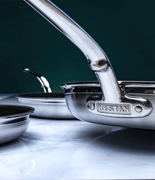 Close-up of stainless steel pans with long handles, placed on a smooth surface. The foreground pan has HESTAN embossed on its handle. The background is a dark, solid color.