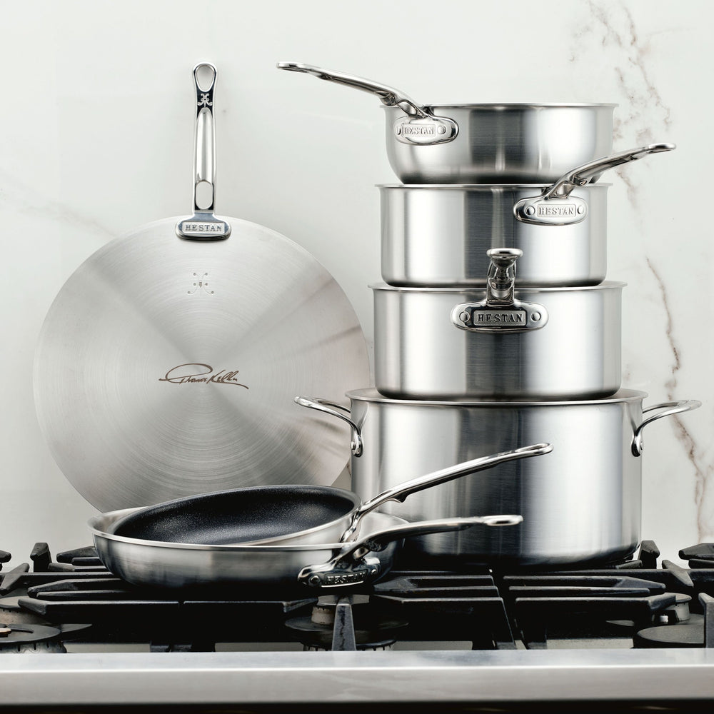 The Thomas Keller Commercial Clad Stainless Steel 7-Piece Cookware Set, including saucepans, a sauté pan, frying pan, and large lid, is displayed on a stovetop with a marble backsplash in the background.