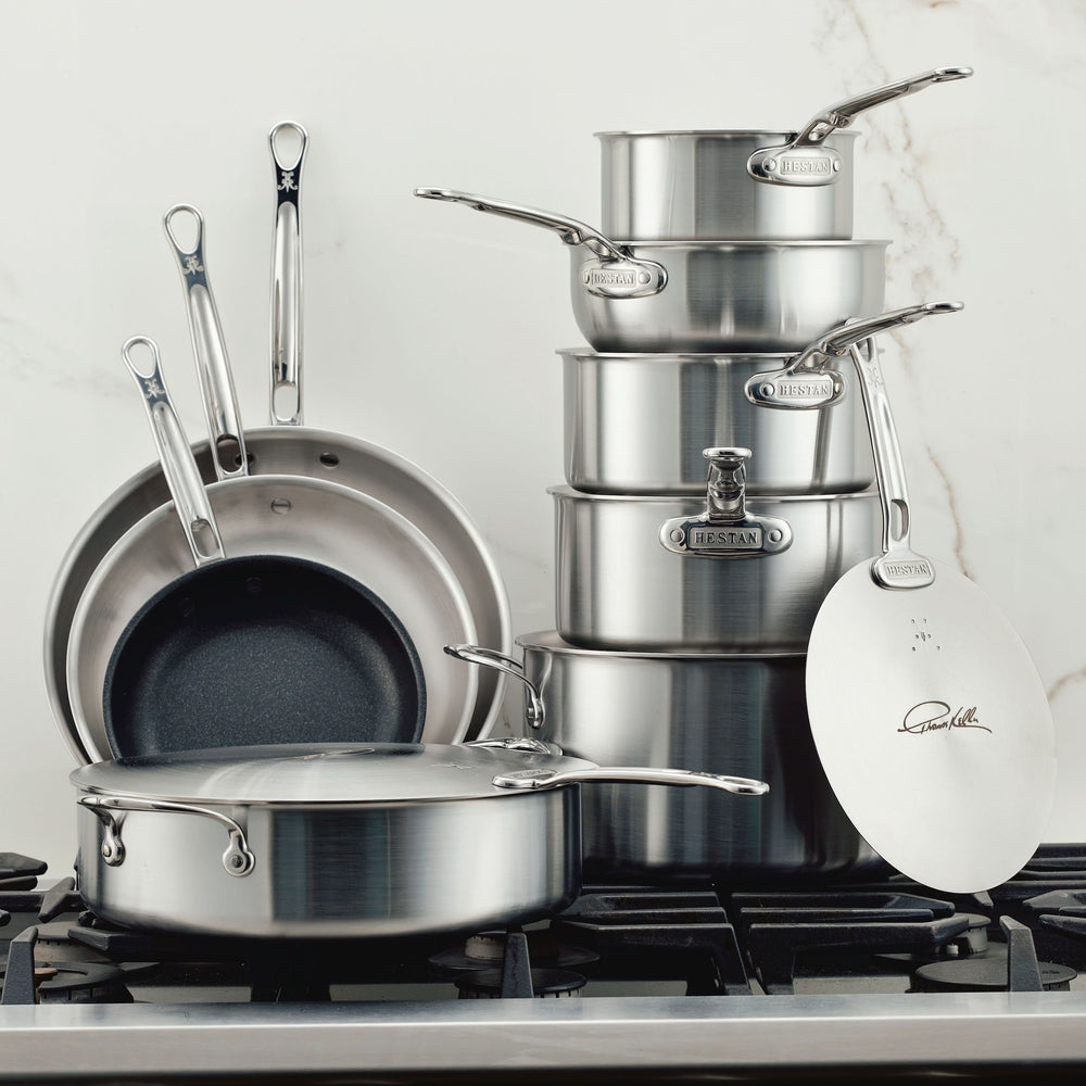 The Thomas Keller Commercial Clad Stainless Steel 11-Piece Cookware Set, featuring pans and pots with lids, sits stacked on a stovetop against a white marble backsplash—ideal for anyone seeking professional-quality cookware.