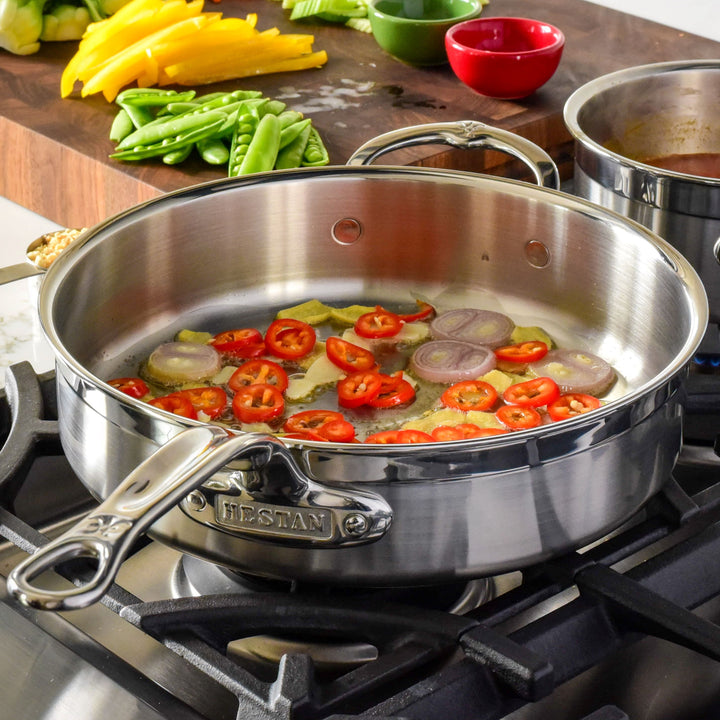 A ProBond® Forged™ Professional Clad Stainless Steel Covered Sauté Pan sizzles with sliced red chili peppers, shallots, and garlic in oil on the stove. On a cutting board are yellow bell pepper and snap peas, with another pot nearby.