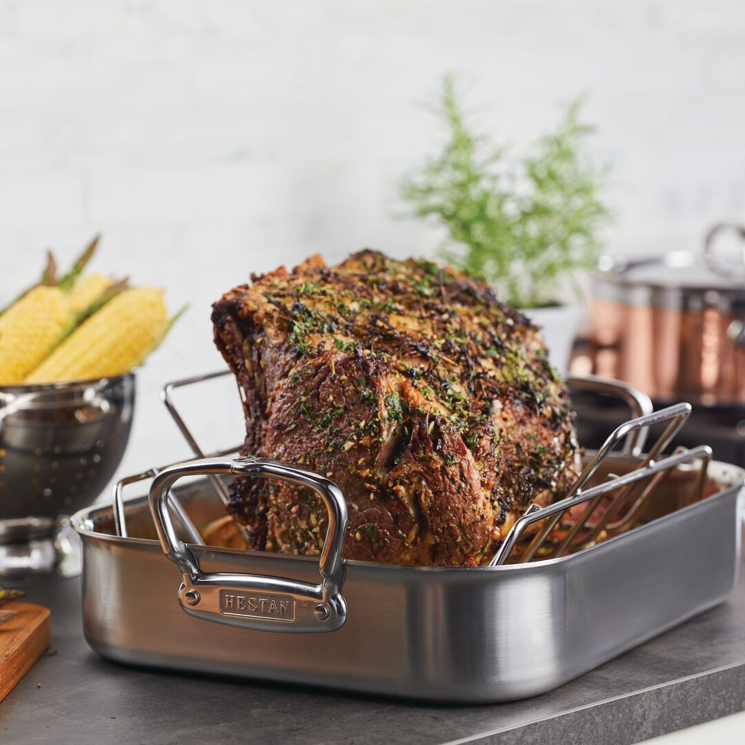 A roast sits in a Hestan Provisions 14.5-inch Classic Clad Roaster with Rack, crusted with herbs. Corn rests in a colander beside a gleaming copper-finished pot, while a sprig of herbs stands proudly in a small container.
