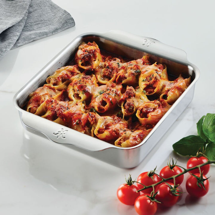 A Hestan OvenBond OvenBond Tri-ply Square Baker holds stuffed pasta shells with tomato sauce and melted cheese, set on a white surface next to fresh tomatoes, basil, and a gray cloth in the background.