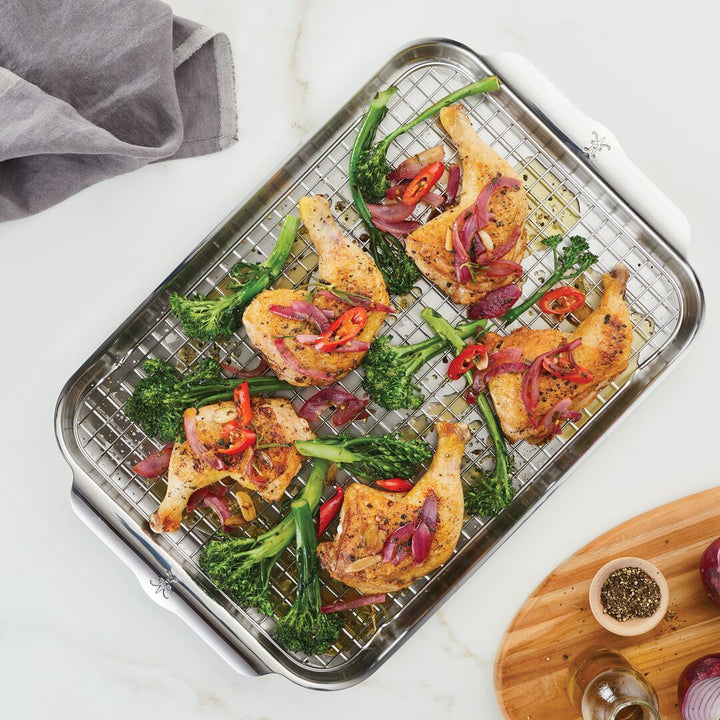 Roasted chicken legs with broccolini, red onions, and sliced red chili peppers rest on the Hestan OvenBond Tri-ply 3-Piece Set rack over a baking tray. A gray napkin and a wooden board with seasonings sit nearby.