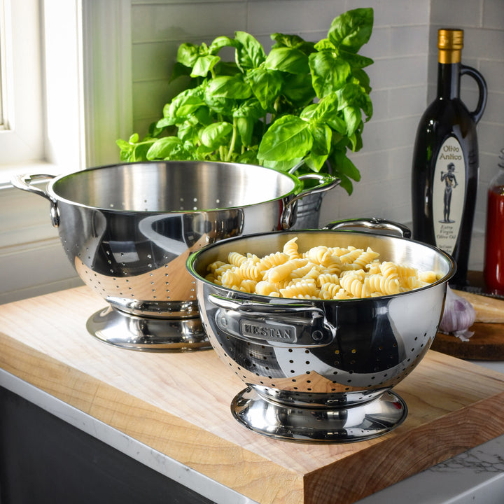 Two Hestan Provisions stainless steel colanders with ergonomic handles rest on a wooden kitchen counter. One is empty, and the other holds cooked pasta. A bottle of olive oil, garlic, and fresh basil are in the background. These dishwasher-safe colanders ensure easy cleaning.