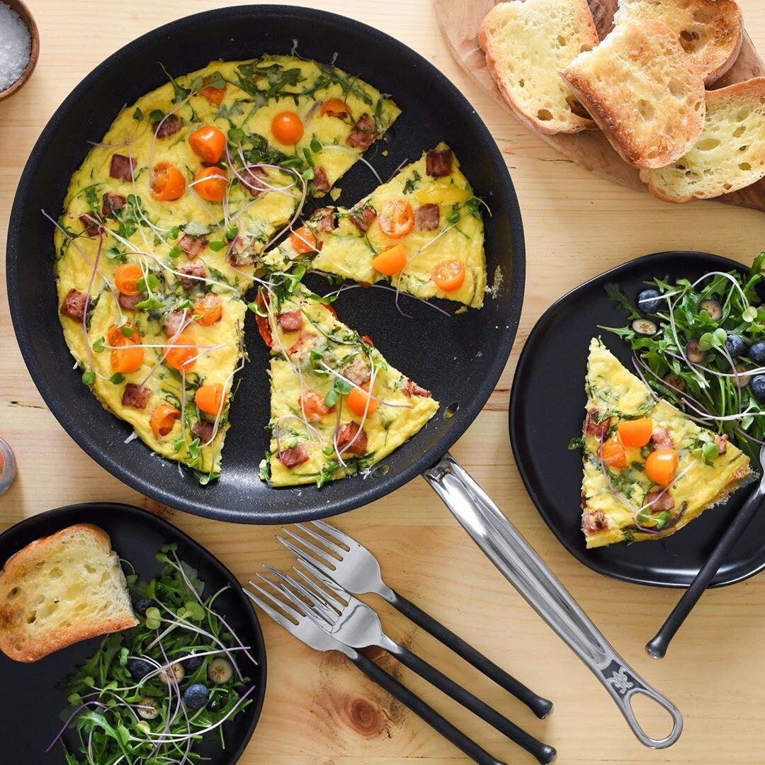 A skillet frittata with cherry tomatoes, greens, and meat is sliced, one piece plated. Toasted bread, microgreens, and utensils sit on a light wooden table beside the ProBond® Forged™ Brushed Clad Stainless Steel TITUM® Nonstick Skillet.