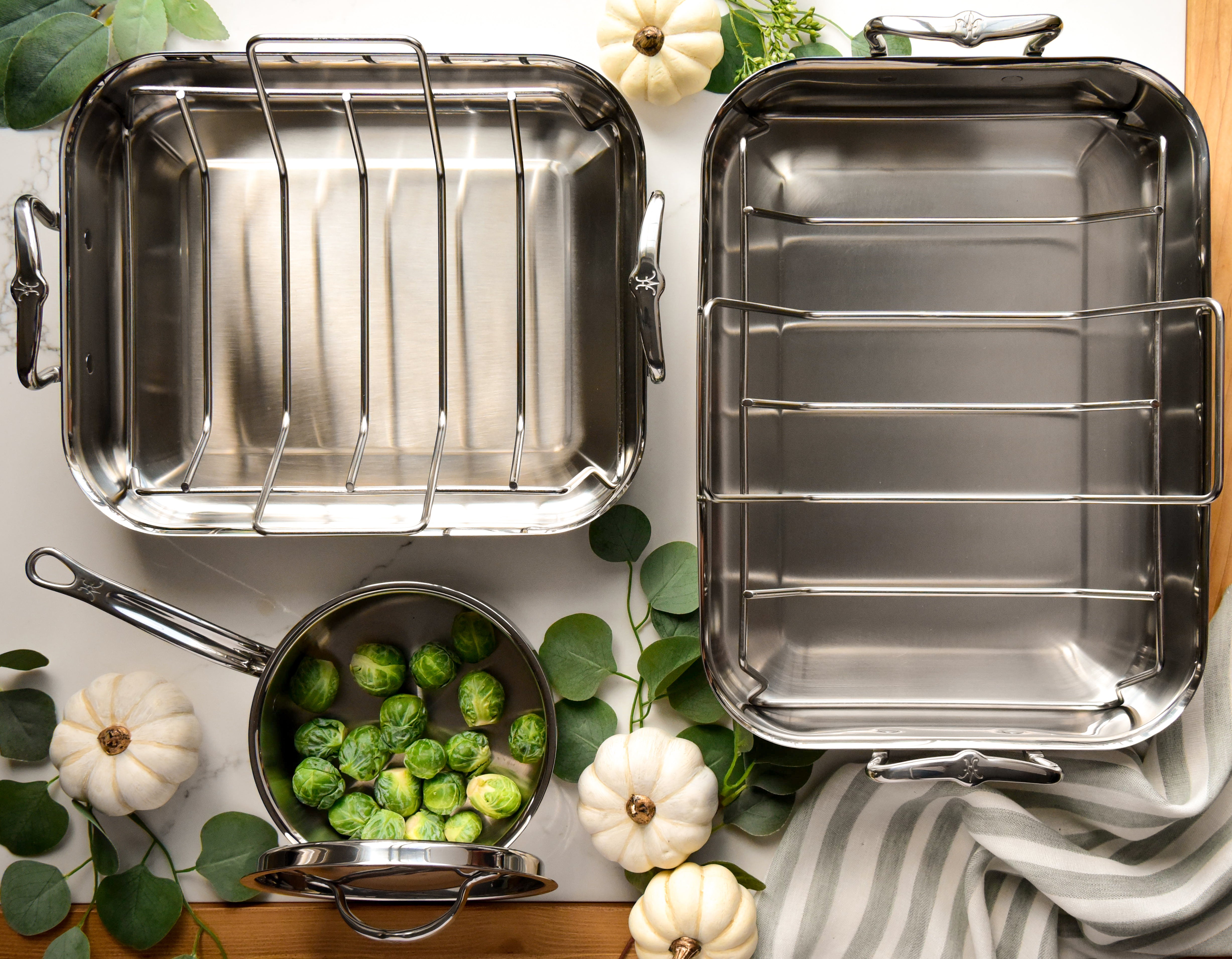 Three Hestan Provisions 16.5-inch Classic Clad Roasters with Racks are elegantly arranged on a surface alongside a saucepan of Brussels sprouts. White pumpkins, green leaves, and a light gray striped cloth enhance the setting, and their superior heat conductivity ensures perfect roasting every time.