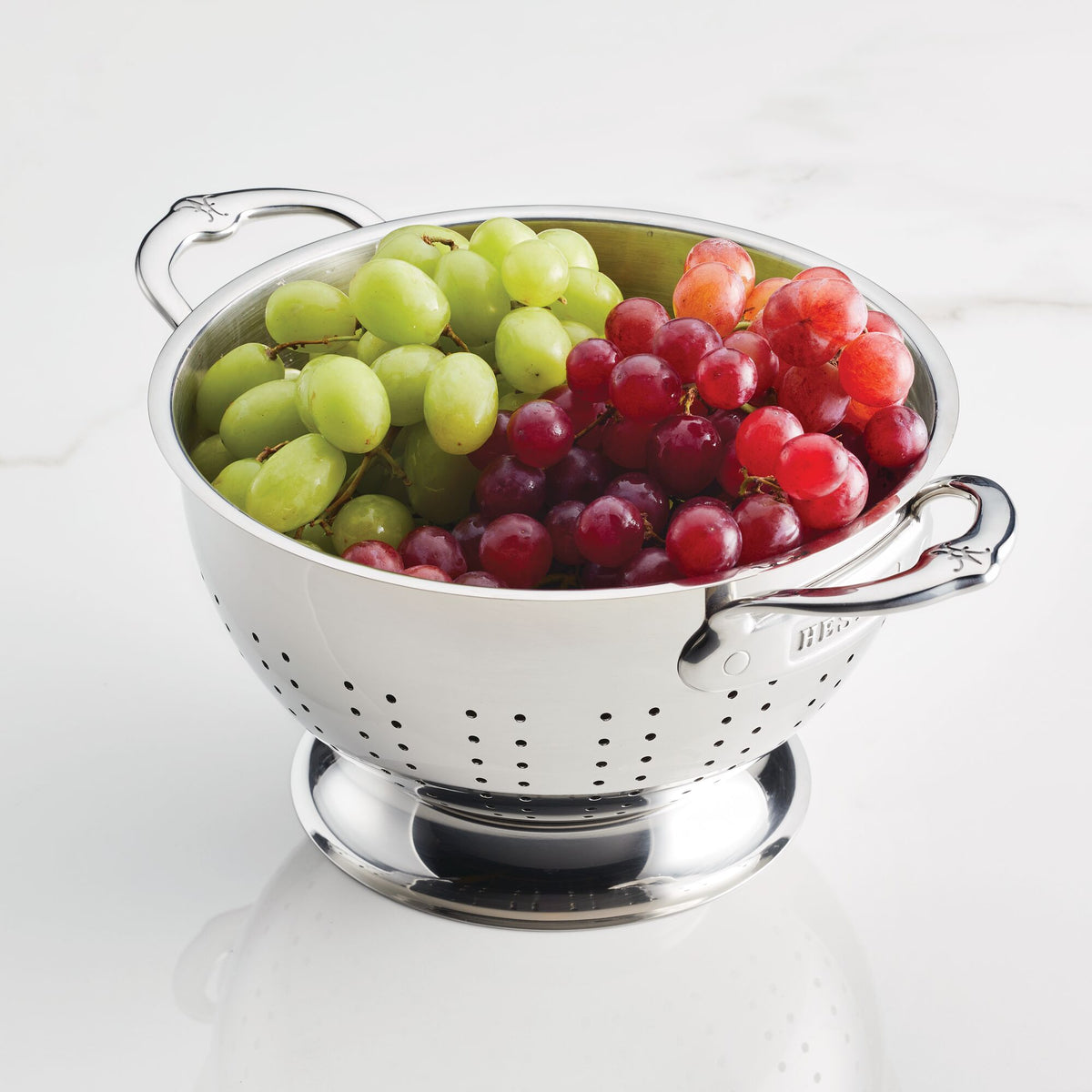 A Hestan Provisions Stainless Steel Colander from the 2-Piece set elegantly holds green grapes on the left and red on the right against a white marble backdrop, with its dual handles framing the scene gracefully.