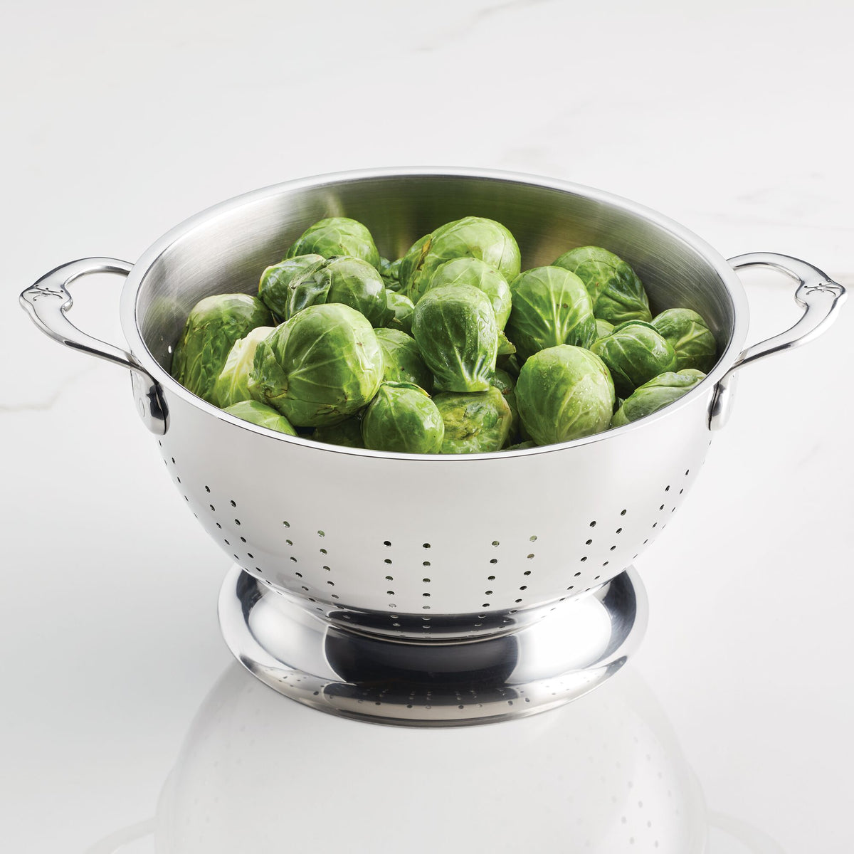 A Hestan Provisions Stainless Steel Colander from the 2-Piece Set rests on a white surface, filled with fresh Brussels sprouts. Its perforated with small holes, boasting ergonomic handles and dishwasher-safe convenience for practicality.