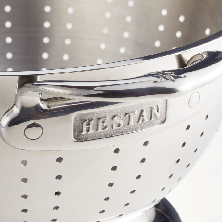 A close-up of a stainless steel colander with perforated holes showcases its handle engraved with Hestan Provisions. Part of a 2-piece set, this polished, dishwasher-safe colander is designed for superior drainage and rests against a neutral background.